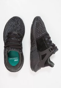 Svarta sportskor med texturerad stickad ovandel, mockadetaljer och gummisulor. Insidan har en grön innersula med "EQUIPMENT"-märkning.