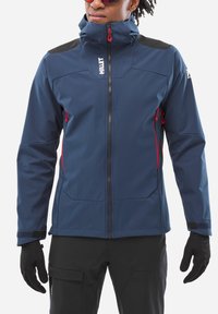 Personne portant une veste d'extérieur zippée bleue avec des tirettes de fermeture éclair rouges, des gants noirs et un pantalon noir sur fond blanc.