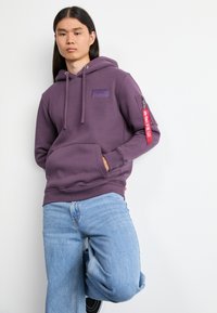 Sudadera de algodón morado con cordones, bolsillo tipo canguro y parche de marca. Cuenta con un bolsillo lateral con cremallera y tirador de cremallera rojo.