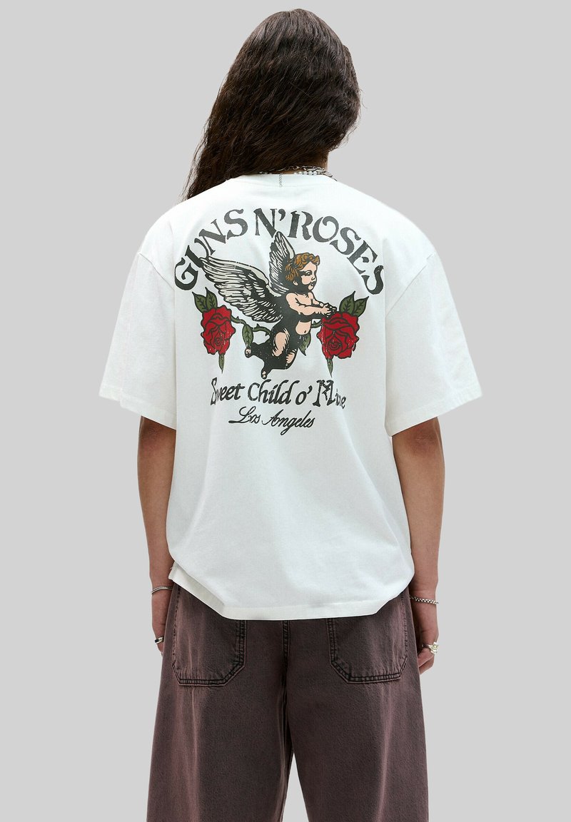 Valkoinen ylisuuri t-paita, jossa on graafinen kuva siipisestä cherubista punaisilla ruusuilla ja teksti "GUNS N' ROSES" sekä "Sweet Child o' Mine" takana.