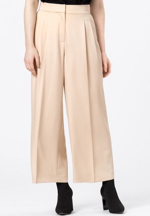 Pantalon beige taille haute à jambes larges porté avec des bottines à talons noires et un haut noir, visible de la taille aux pieds.