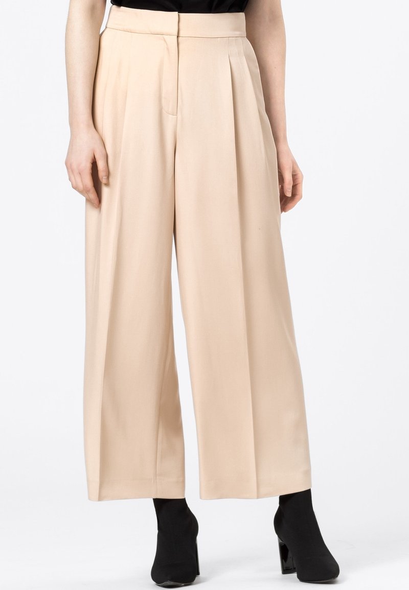 Pantalon beige taille haute à jambes larges porté avec des bottines à talons noires et un haut noir, visible de la taille aux pieds.