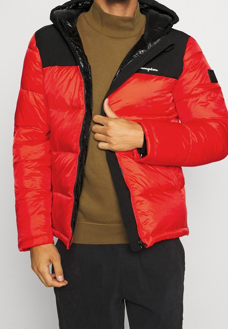 Homme portant une doudoune rouge et noire par-dessus un col roulé marron, dézippant partiellement la veste d'une main.