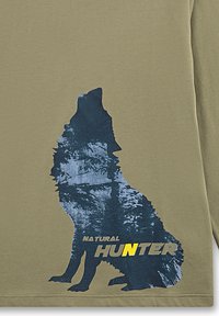 Olijfgroene lange mouw shirt met een donkerblauwe wolfsilhouette met een bospatroon, en "NATURAL HUNTER" in gele tekst onderaan.