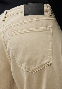 Πίσω τσέπη μπεζ τζιν με ορατή ραφή και μαύρη δερμάτινη ετικέτα επωνυμίας ραμμένη στη ζώνη μέσης.