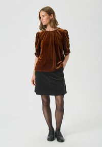 Bruine fluwelen top met opstaande mouwen, gecombineerd met een zwarte rok tot de knie. De outfit heeft een ronde halslijn en een soepele textuur.