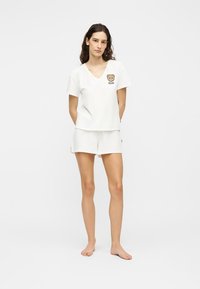 Γυναίκα όρθια, ξυπόλυτη, που φοράει λευκό T-shirt με V-λαιμόκοψη και λογότυπο αρκουδάκι, καθώς και λευκά σορτς, μπροστά σε μονόχρωμο φόντο.