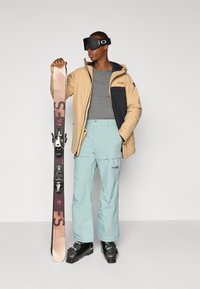 Jachetă de schi bej și neagră, cămașă gri, pantaloni de zăpadă bleu deschis, ochelari de schi negri și bocanci, ținând un schi cu prinderi.