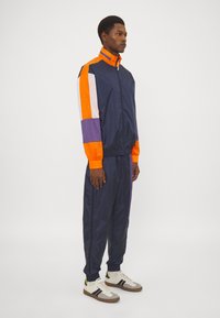 Marine vindjakke med oransje, lilla og hvite fargeblokker; elastiske mansjetter; matchende bukser; komplementert av grå og hvite joggesko med svarte striper.