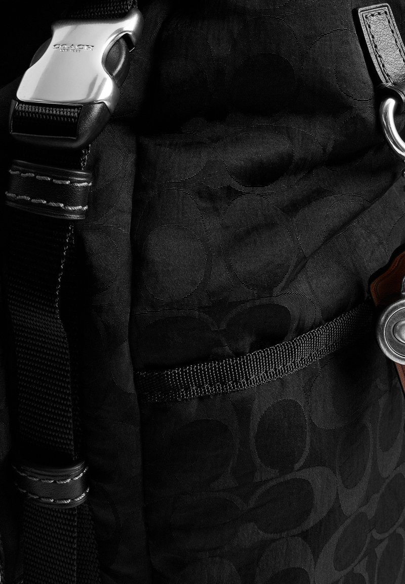 Black fabric bag with subtle circular pattern, black strap with silver buckle engraved "COACH New York," and silver keyring attached.
