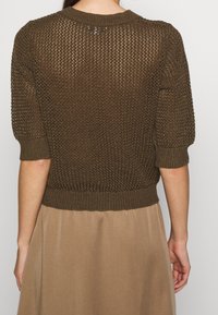 Bruine gebreide trui met korte pofmouwen, voorzien van een geribde zoom en textuurpatroon, gedragen over een lichtbruine jurk.