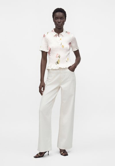 Femme debout, main dans la poche, portant un pantalon blanc à jambes larges, un chemisier blanc à manches courtes à motif floral avec un ourlet à volants, et des sandales à talons noires.