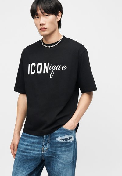 Νεαρός άνδρας που φοράει μαύρο κοντομάνικο μπλουζάκι με την επιγραφή "ICONique", μπλε σκισμένο τζιν και κολιέ με χάντρες, όρθιος με το ένα χέρι στην τσέπη.