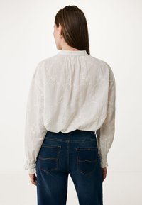 Witte blouse met lange mouwen, versierd met bloemenborduurwerk en gerimpelde manchetten, gecombineerd met donkerblauwe high-waisted jeans met achterzakken.