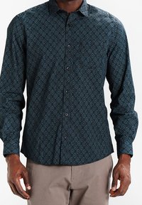 Langsleeve shirt in donkerblauwe stof met een geometrisch patroon. Kenmerkt zich door een puntkraag, knoopsluiting en een getailleerde pasvorm.