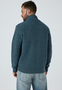 Tumši teļa zaļš adīts džemperis ar augstu apkakli un ribotām piedurknēm, ar strukturētu rakstu un brīvu piegriezumu. Valkāts kopā ar gaiši zilām džinsām.