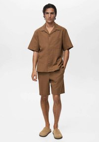 Cămașă maro cu mânecă scurtă, cu nasturi, dotată cu un buzunar aplicat, asortată cu pantaloni scurți și pantofi slip-on bej. Textură netedă, croială lejeră.