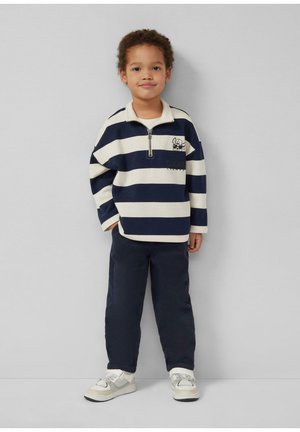 Junges Kind steht vor einer schlichten Wand und trägt einen blau-weiß gestreiften Pullover, blaue Hosen und weiße Turnschuhe.