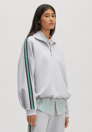 Frau trägt ein hellgraues Sweatshirt mit halbem Reißverschluss und gestreiftem Besatz an den Ärmeln, darüber eine grün-rosa gestreifte Hemdbluse und passende Hose.