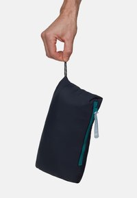 Pochette en tissu bleu marine tenue à la main, dotée d'une fermeture éclair turquoise et d'une surface texturée. Comprend un onglet de tirage avec des marques de mesure.