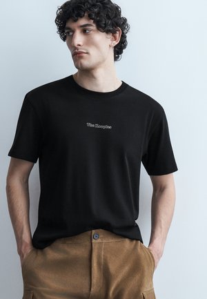 Jeune homme aux cheveux bouclés portant un t-shirt noir "The Kooples" et un pantalon marron, les mains dans les poches, regardant vers la gauche sur un fond uni.