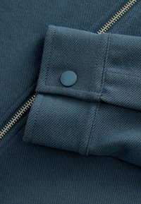 Stof van een blauwgroene jas met een geribbelde textuur, voorzien van een ritssluiting en een ronde knoop op de manchetten. Gouden ritssluiting accenten.