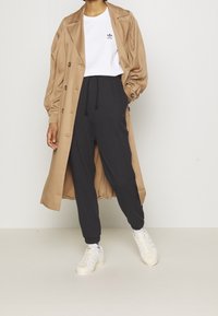 Henkilö, joka käyttää vaaleanbeigeä sadetakkia, valkoista Adidas-t-paitaa, mustia jogginghousuja, valkoisia sukkia ja valkoisia Adidas-tennareita, seisoo yksinkertaista taustaa vasten.