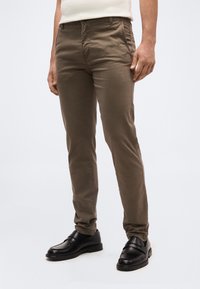 Άνδρας που φοράει καφέ παντελόνια στενής εφαρμογής και μαύρα δερμάτινα μοκασίνια, στέκεται μπροστά σε ένα απλό φωτεινό φόντο.