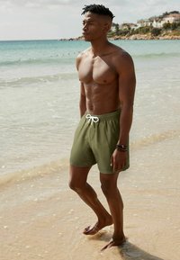 Rövid zöld fürdőnadrág fehér zsinórral, egy homokos tengerparton álló személyen, vízzel a háttérben, amely laza fazont és sima textúrát mutat be.