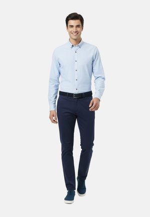 Hombre caminando hacia adelante con una camisa de botones azul claro, pantalones azul oscuro con un cinturón negro y zapatillas deportivas azules, sonriendo.