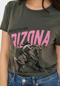 Camiseta verde oliva con un estampado gráfico de un vaquero montado a caballo, con "ARIZONA" en rosa y el texto "RODEO COWGIRL" arriba.