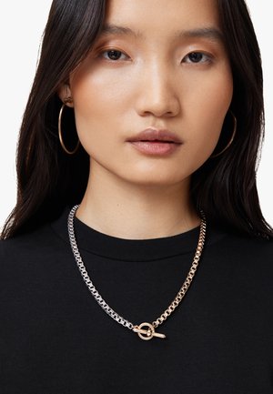 Collana in metallo argento e oro con chiusura a patta rotonda, indossata su una maglietta nera con scollo a barco.