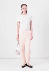 Bílá tričko, světle růžové tepláky a černé baleríny. Modelka drží malou šedou kabelku na rameno. Jednoduchý design s elastickým pasem.