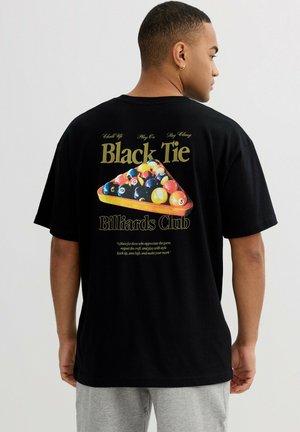 Hombre con camiseta negra con bolas de billar de colores en soporte triangular y texto "Black Tie Billiards Club" en la espalda, pantalones grises.