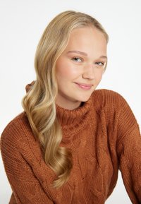 Knitattava kaulahuivi lämpimässä ruskeassa värissä, jossa on teksturoitu palmikkokuvio. Mallilla on pitkät vaaleat hiukset, jotka valuavat olkapään yli, hymyillen lempeästi.