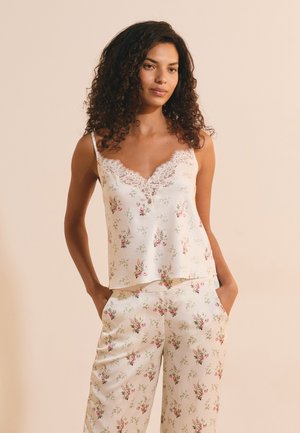 Donna con capelli ricci che indossa una canottiera color crema rifinita con pizzo floreale e pantaloni a gamba larga abbinate, con le mani nelle tasche su sfondo beige.