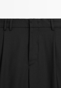Pantalons noirs avec une taille plate, des passants de ceinture et une texture lisse. Comprend une poche unique et un design minimaliste, mis en valeur sur un fond blanc.