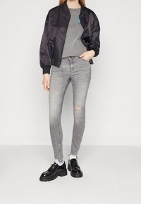 Chaqueta bomber negra de satén, camiseta gris, jeans skinny gris rasgados y zapatos gruesos negros con un acabado brillante. Sin patrones o acentos visibles.