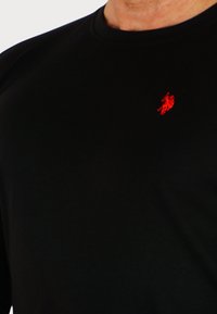 Zwarte T-shirt van katoen, met een ronde hals en korte mouwen. Opvallend rood geborduurd logo aan de linkerkant.