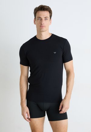 Schwarzes, eng anliegendes T-Shirt aus glattem Stoff, mit einem kleinen Logo auf der Brust. Das Modell trägt schwarze Shorts und steht vor einem schlichten Hintergrund.