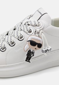 Zapatilla blanca de cuero con cordones estampados, ojales plateados y un charm colorido que representa una figura estilizada. Logo "KARL LAGERFELD" presente.