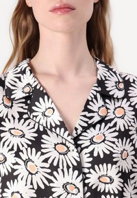 Maglia da pigiama floreale in bianco e nero con colletto. Presenta margherite con centri arancioni e una consistenza del tessuto morbida. Chiusura a bottoni sullo scollo.