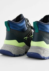 Wysokie sportowe sneakersy z czarno-zielonego materiału syntetycznego, niebieskimi akcentami, teksturowaną szaro-skalną podeszwą i odblaskowymi detalami.