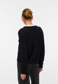 Femme aux cheveux blonds attachés en chignon, portant un pull noir et un jean foncé, debout, dos au spectateur, sur un fond uni.
