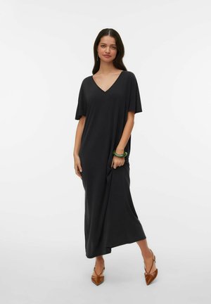 Femme portant une robe maxi noire ample à col en V et manches courtes, des chaussures pointues marron et un bracelet vert, debout sur un fond clair.