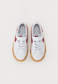 Bela Nike teka z rdečim logotipom, gumijastim podplatom, teksturiranim tekstilnim podlogo in vezalnim dizajnom, ki ima dodatno zanko.