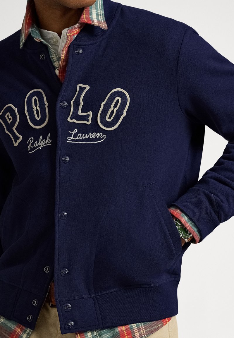 Navy blue jersey cardigan with cream embroidered letters "POLO" and "Ralph Lauren." Features snap buttons and side pockets, layered over a plaid shirt.