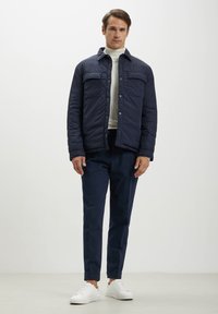 Giacca trapuntata blu navy con due tasche sul petto, abbinata a pantaloni scuri e sneaker bianche. Tessuto morbido, apertura con bottoni, design del colletto classico.