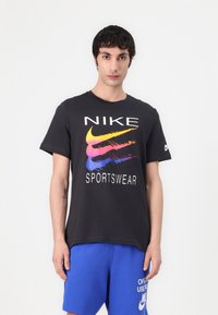 Tricou din bumbac negru cu text alb "NIKE" și grafică colorată a swoosh-ului. Modelul este asortat cu pantaloni scurți albaștri. Guler rotund simplu.