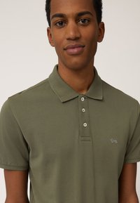 Polo de color verde oliva hecho de una tela texturizada, con collar, tres botones y un pequeño logo bordado en el pecho.